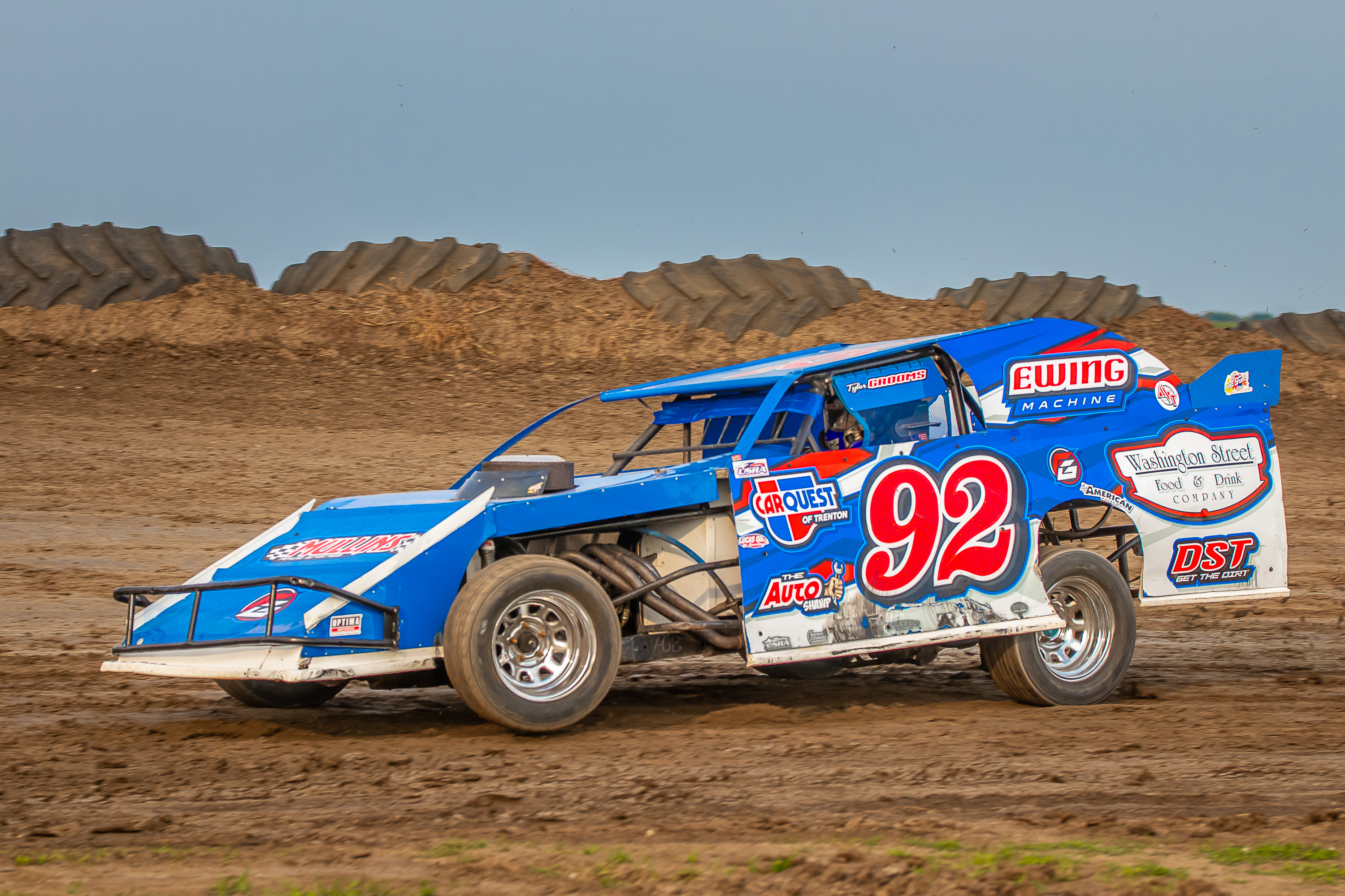 Blue and white dirt track race car number 92 racing on a dirt oval with DST and sponsor logos.